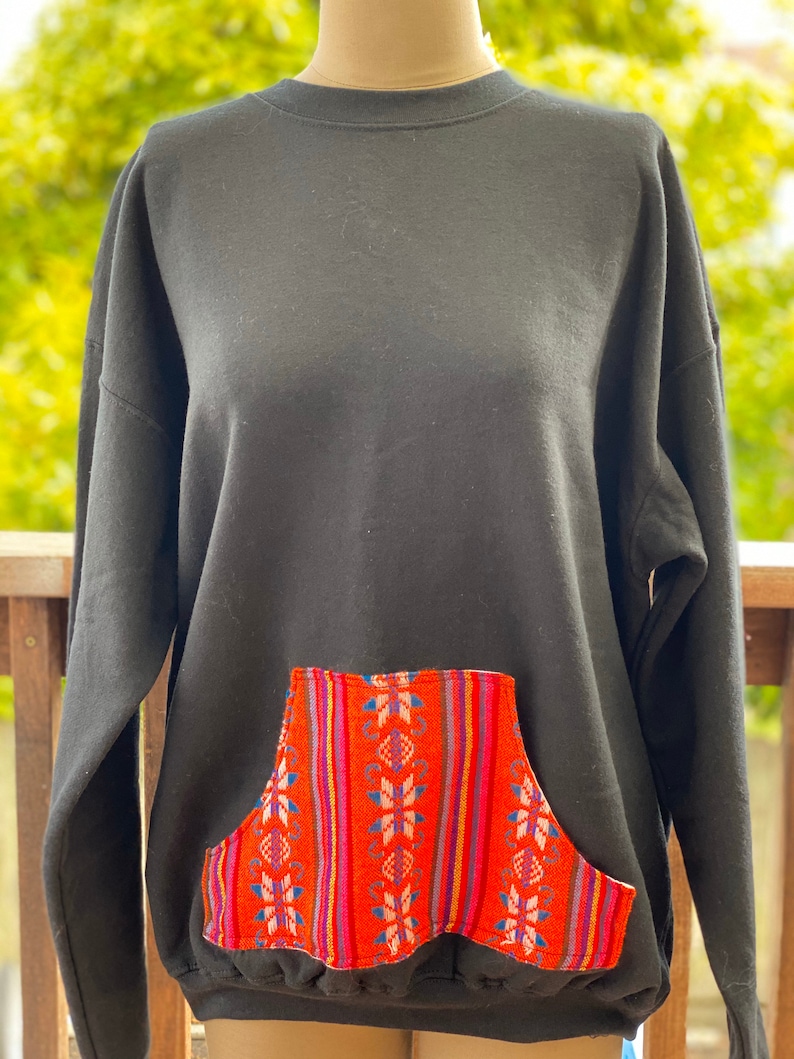 May include: Black sweatshirt with a large, colorful, patterned pocket on the front. The pocket has a geometric design with red, pink, blue, and white stripes.