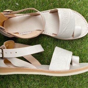 May include: White leather sandals with a buckle closure and a decorative embossed design. The sandals have a white sole and a comfortable strap.