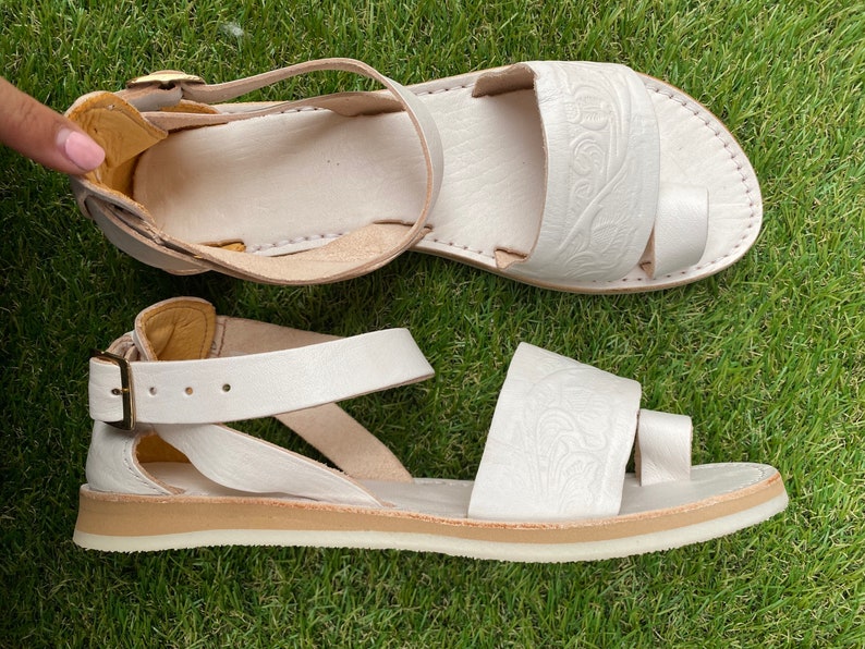 May include: White leather sandals with a buckle closure and a decorative embossed design. The sandals have a white sole and a comfortable strap.