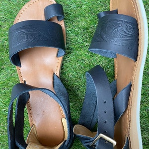 May include: A pair of black leather sandals with an ankle strap and a toe loop. The sandals have a floral design and a brown sole.