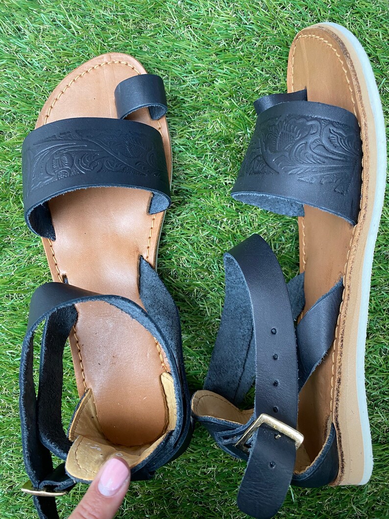 May include: A pair of black leather sandals with an ankle strap and a toe loop. The sandals have a floral design and a brown sole.