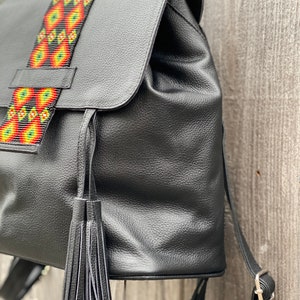 Op de afbeelding: Zwarte leren rugzak met een kleurrijke kralenband en een zwarte kwast. De rugzak heeft een klep sluiting en een ritsvak aan de voorkant.