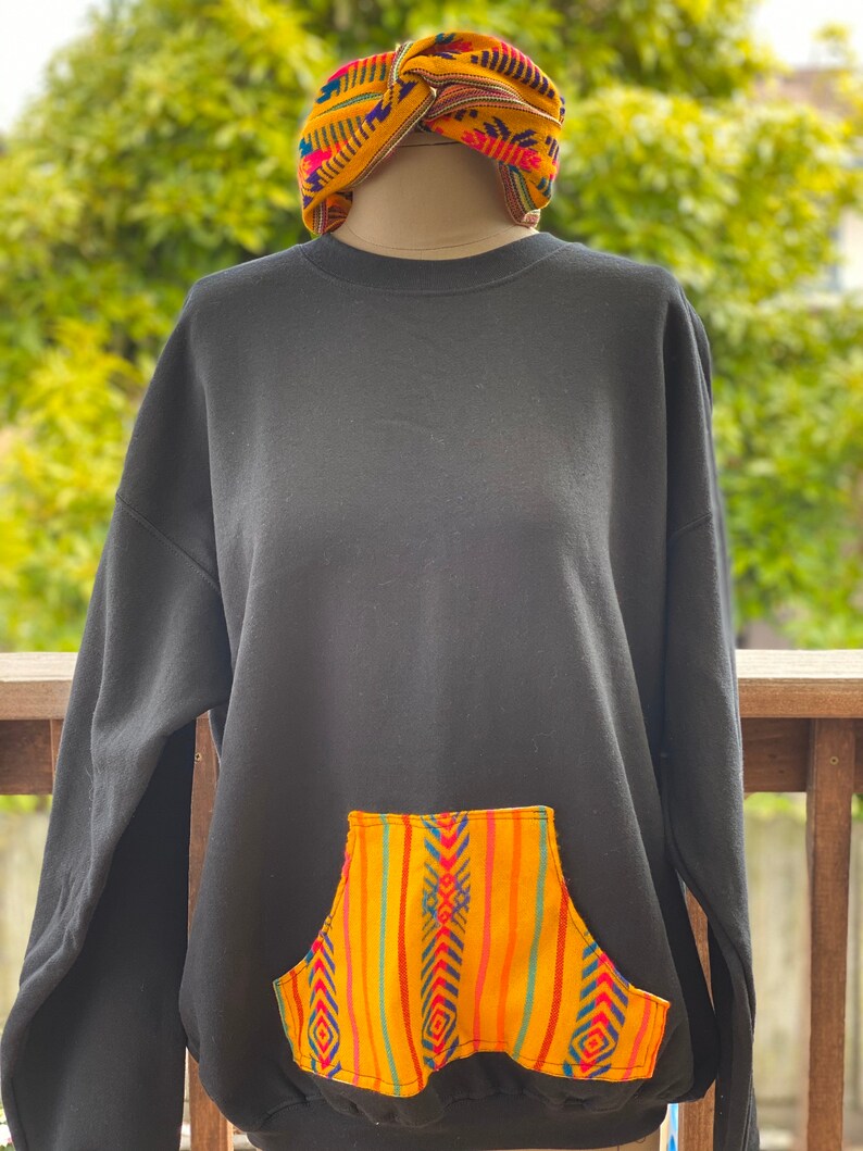 May include: Black sweatshirt with a large pocket on the front. The pocket is made of colorful fabric with a geometric pattern in yellow, orange, red, blue, and green.