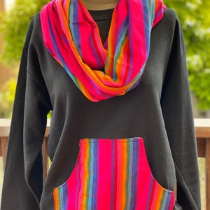 May include: Black sweatshirt with a large pocket on the front. The pocket is lined with a colorful striped fabric. The sweatshirt also has a colorful striped scarf around the neck.