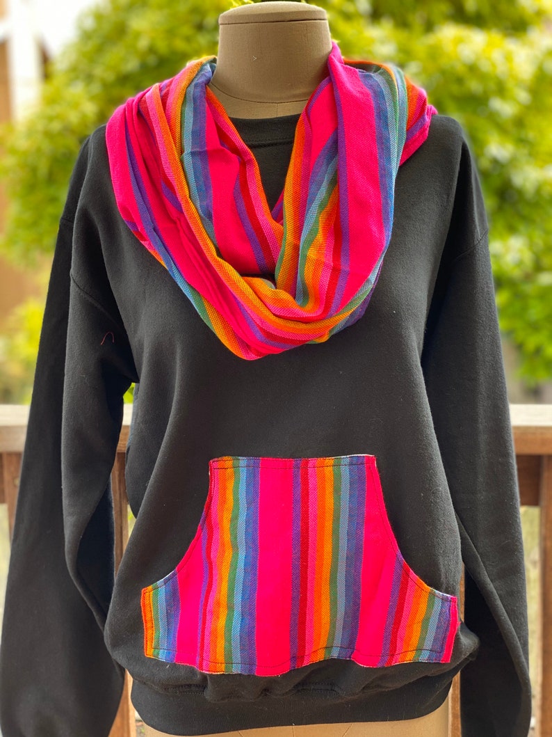 May include: Black sweatshirt with a large pocket on the front. The pocket is lined with a colorful striped fabric. The sweatshirt also has a colorful striped scarf around the neck.