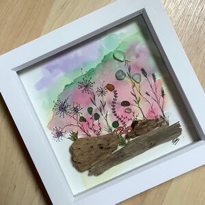 Può includere: Un'opera d'arte ad acquerello incorniciata con un paesaggio colorato con uno sfondo rosa, verde e viola. L'opera d'arte include fiori dipinti, piccole pietre e pezzi di legno trasportati dalla corrente, il tutto racchiuso in una cornice bianca.