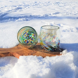 May include: Two colourful glass tumblers with a speckled and swirled design sit on a wooden chopping board in the snow.