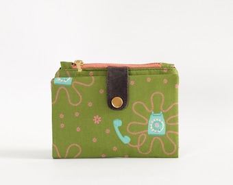 Può includere: Un portafoglio in tessuto verde con un motivo floreale e un design di telefono vintage. Il portafoglio ha una patta in pelle marrone con un bottone dorato.