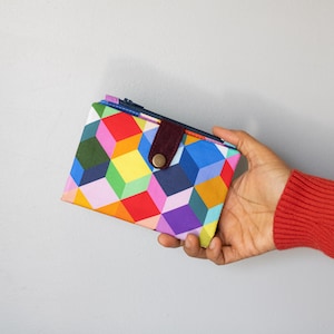 May include: A wallet with a vibrant geometric cube design in red, yellow, blue, green, and purple. It features a dark brown strap with a gold button clasp. The wallet is held in a person's hand.