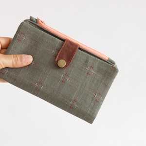 Könnte beinhalten: Eine rechteckige, olivgrüne Geldbörse mit einem rosa Reißverschluss und einem braunen Leder-Druckknopfverschluss. Die Geldbörse hat ein Kreuzstichmuster in Rot und Weiß. Die Geldbörse wird in einer Hand gehalten.