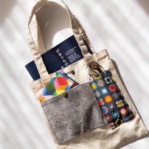 May include: Beige canvas tote bag with a variety of items inside. The bag has a front pocket with a gray speckled fabric and a floral print pocket. Inside the bag are books and a colorful geometric patterned wallet.