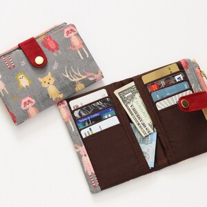 May include: Two fabric wallets, one with a gray background and a colorful animal print, and the other with a brown interior and a pink and gray animal print. Both wallets have a zipper closure and a red button.