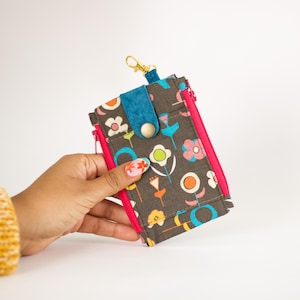 May include: A floral print wallet with a teal accent and a pink zipper. The wallet has a gold-colored clasp and a snap closure. The wallet is held by a person.