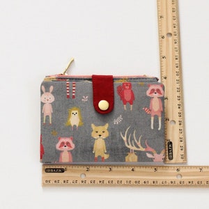 May include: A gray fabric wallet with a red button closure and a zipper. The wallet has a woodland animal print with a fox, deer, raccoon, hedgehog, and bunny. The wallet measures approximately 3.5 inches wide and 4.5 inches tall.