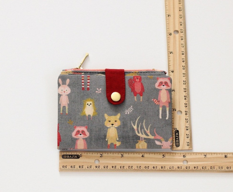 May include: A gray fabric wallet with a red button closure and a zipper. The wallet has a woodland animal print with a fox, deer, raccoon, hedgehog, and bunny. The wallet measures approximately 3.5 inches wide and 4.5 inches tall.