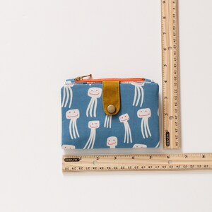 May include: A blue fabric wallet with a zipper closure and a flap with a button. The wallet has a pattern of white smiling jellyfish on a blue background. The flap is a mustard yellow color.