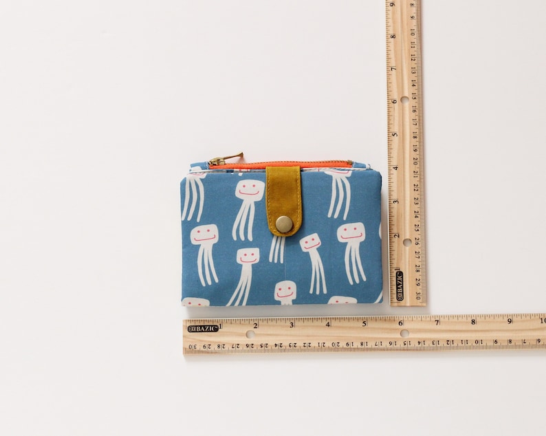 May include: A blue fabric wallet with a zipper closure and a flap with a button. The wallet has a pattern of white smiling jellyfish on a blue background. The flap is a mustard yellow color.