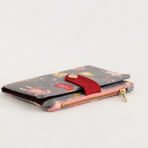 May include: A gray wallet with a pink and red zipper and a red button closure. The wallet has a colorful pattern of animals and flowers.