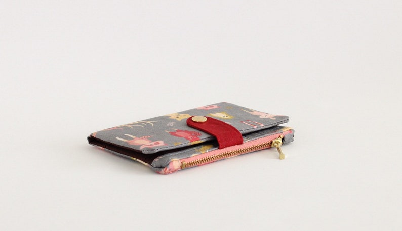May include: A gray wallet with a pink and red zipper and a red button closure. The wallet has a colorful pattern of animals and flowers.