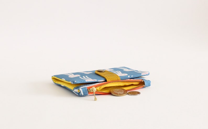 May include: A small blue zippered wallet with a yellow flap and a white pattern of rabbits. The wallet is open and has a few coins inside.