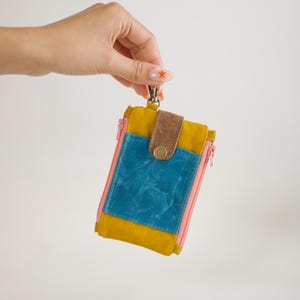 May include: A colourful wallet with a zip closure. The wallet features a mustard yellow, teal, and pink colour scheme, with a brown leather flap secured by a brass button. A silver clasp is attached to the top.