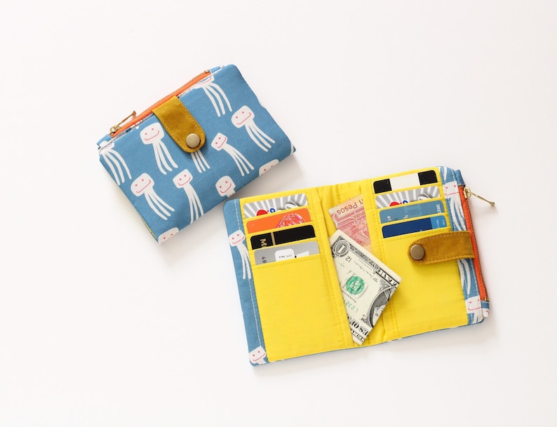 May include: Two fabric wallets, one blue with a smiling jellyfish pattern and one yellow with multiple card slots and a bill compartment. Both wallets have a gold zipper and a gold button.