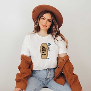May include: A white t-shirt with a graphic of a skeleton in a coffin with the text "I'll sleep when I'm dead". The model is wearing a brown hat and a brown jacket.