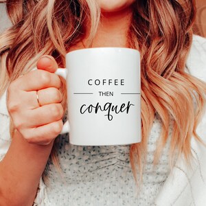 以下が含まれることがあります： 白いセラミック製のコーヒーカップに、黒い文字で「Coffee then conquer」と書かれています。