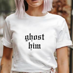 Puede incluir: Camiseta blanca con la frase "ghost him" impresa en fuente gótica negra. La camiseta tiene mangas cortas y un corte relajado.