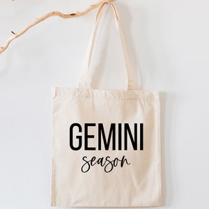 May include: A beige canvas tote bag with black text that reads "GEMINI season".