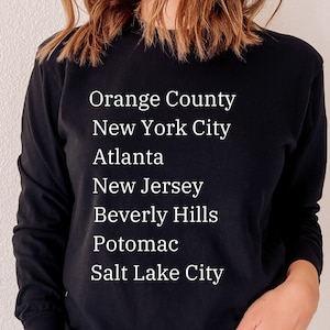 May include: Black long-sleeve shirt with white text listing cities: Orange County, New York City, Atlanta, New Jersey, Beverly Hills, Potomac, and Salt Lake City. The shirt is a solid color.