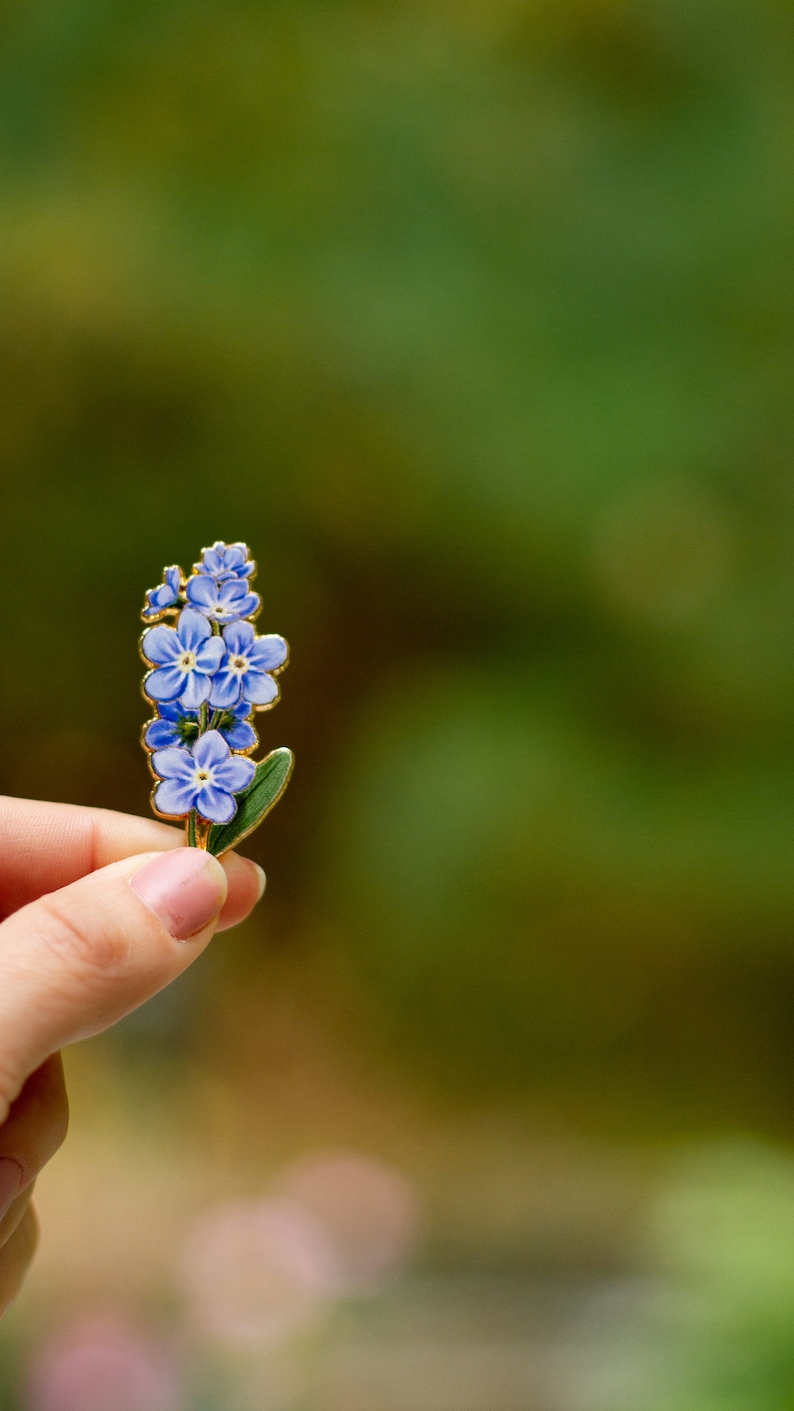 May include: A gold-toned enamel pin featuring a cluster of blue forget-me-not flowers with green leaves.