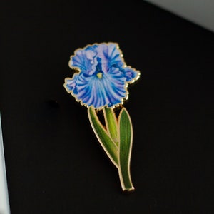 May include: A gold-toned enamel pin featuring a blue and purple iris flower with green leaves.