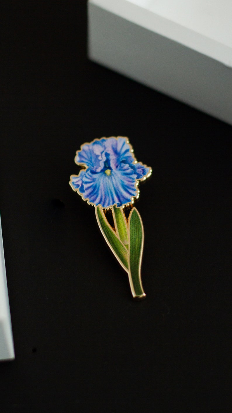 May include: A gold-toned enamel pin featuring a blue and purple iris flower with green leaves.