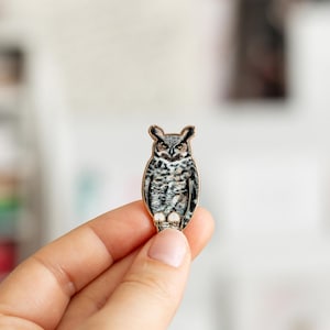 Puede incluir: Un pequeño broche de búho con borde dorado. El búho presenta tonos grises, negros y blancos. El broche se sostiene en una mano, resaltando el diseño detallado. Un accesorio decorativo.