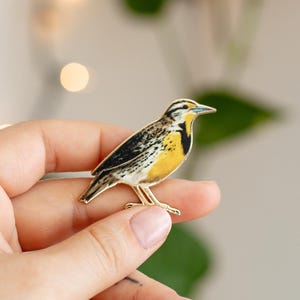 May include: A metal enamel pin shaped like a meadowlark bird. The bird features a bright yellow chest with a black V-shaped patch, brown and white mottled wings, and a slender beak. The pin has a gold-toned metal outline and visible metal legs.