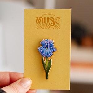 May include: A gold-toned enamel pin featuring a blue iris flower with green leaves. The pin is on a yellow card with the text "The Gray Muse" in a stylized font.