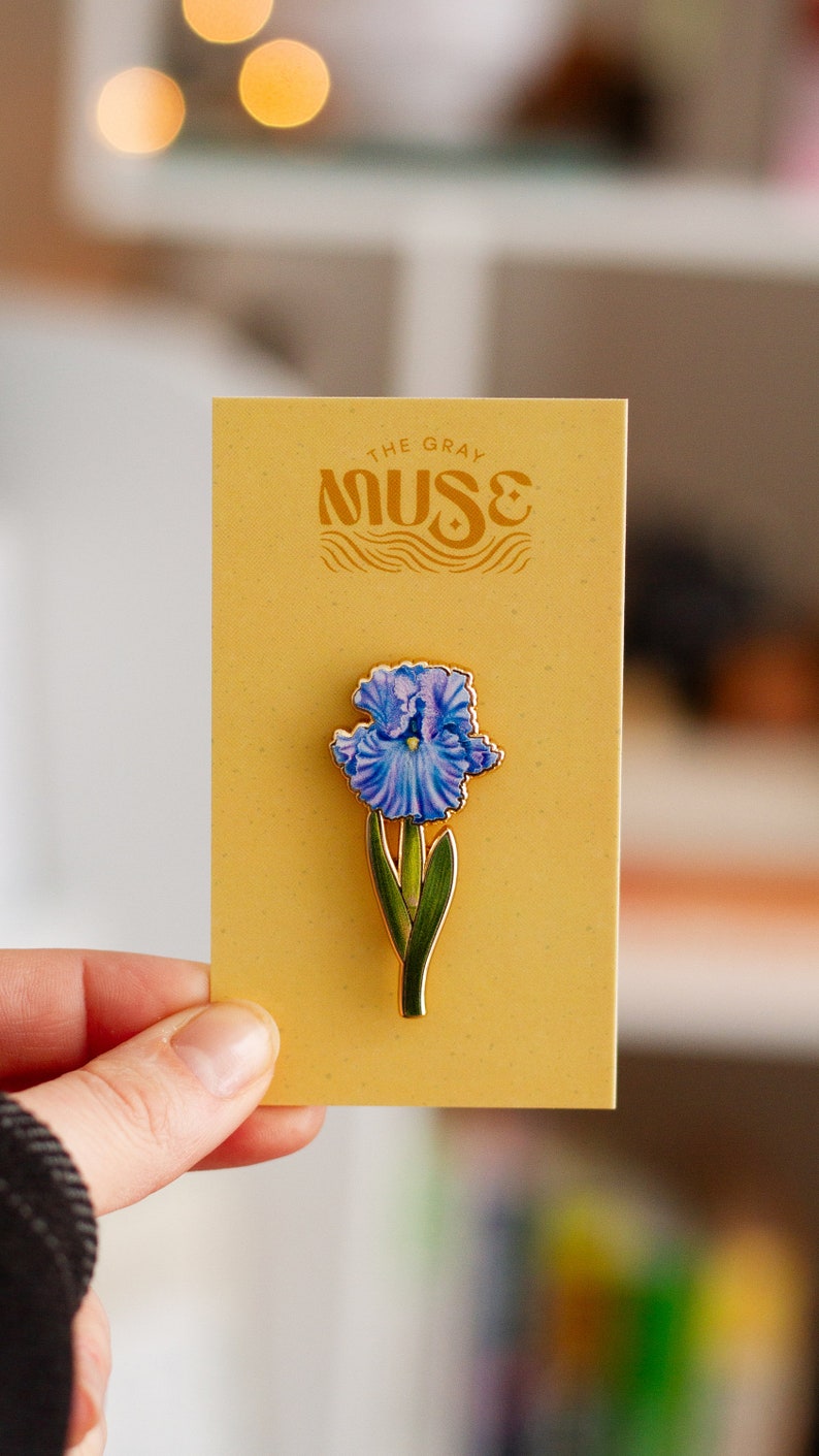 May include: A gold-toned enamel pin featuring a blue iris flower with green leaves. The pin is on a yellow card with the text "The Gray Muse" in a stylized font.
