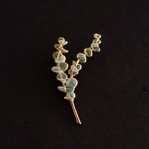 May include: A gold-coloured metal brooch shaped like a eucalyptus branch. The leaves are a muted green with gold edging, and the stem is a warm gold colour. The brooch is set against a black background.