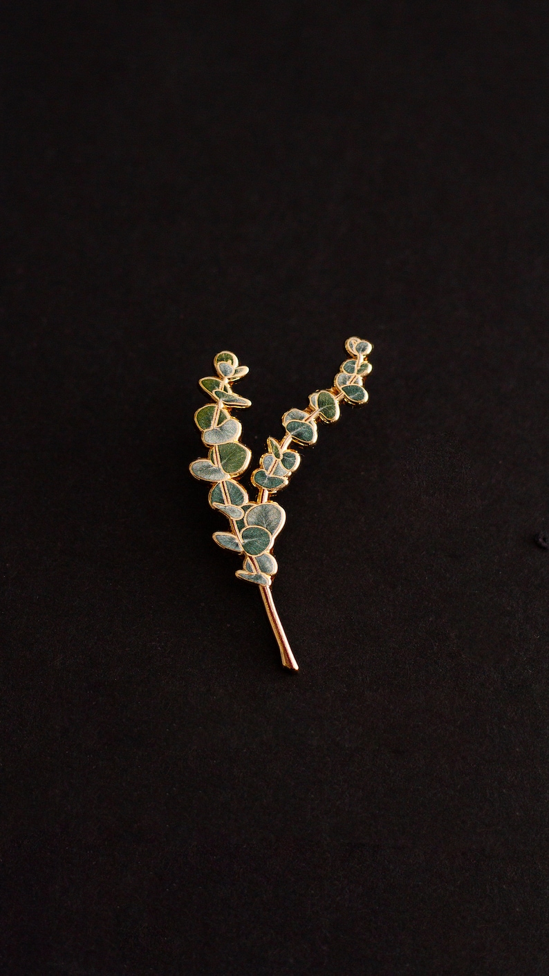May include: A gold-coloured metal brooch shaped like a eucalyptus branch. The leaves are a muted green with gold edging, and the stem is a warm gold colour. The brooch is set against a black background.