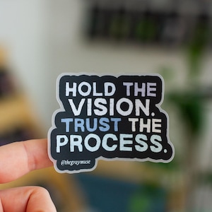 May include: A black sticker with white and light blue text that reads "HOLD THE VISION. TRUST THE PROCESS." The sticker has a white border and the text "@thegraymuse" at the bottom. The sticker is held between two fingers with pink nail polish.