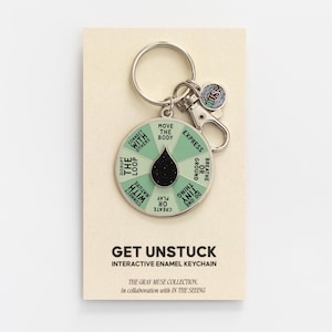 May include: A round, light green enamel keyring featuring a spinning wheel with text prompts including MOVE THE BODY, EXPRESS, BREATHE OR GROUND, DO ONE TINY THING, PLAY OR CREATE, CONNECT WITH NATURE, DISRUPT THE LOOP, and CONNECT WITH OTHERS.