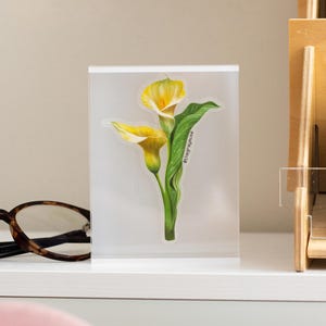 May include: A clear acrylic block featuring a detailed illustration of two yellow calla lilies with green leaves. The artwork includes the text "@thegraymuse" and is set against a neutral background, suggesting a decorative item or print.