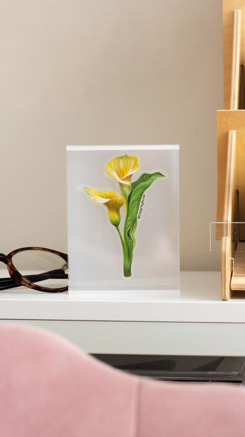 May include: A clear acrylic block featuring a detailed illustration of two yellow calla lilies with green leaves. The artwork includes the text "@thegraymuse" and is set against a neutral background, suggesting a decorative item or print.