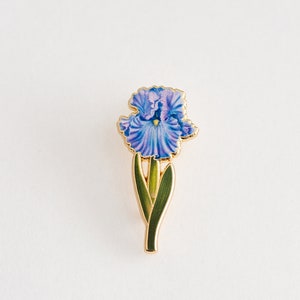 May include: A gold-toned enamel pin featuring a purple iris flower with green leaves.