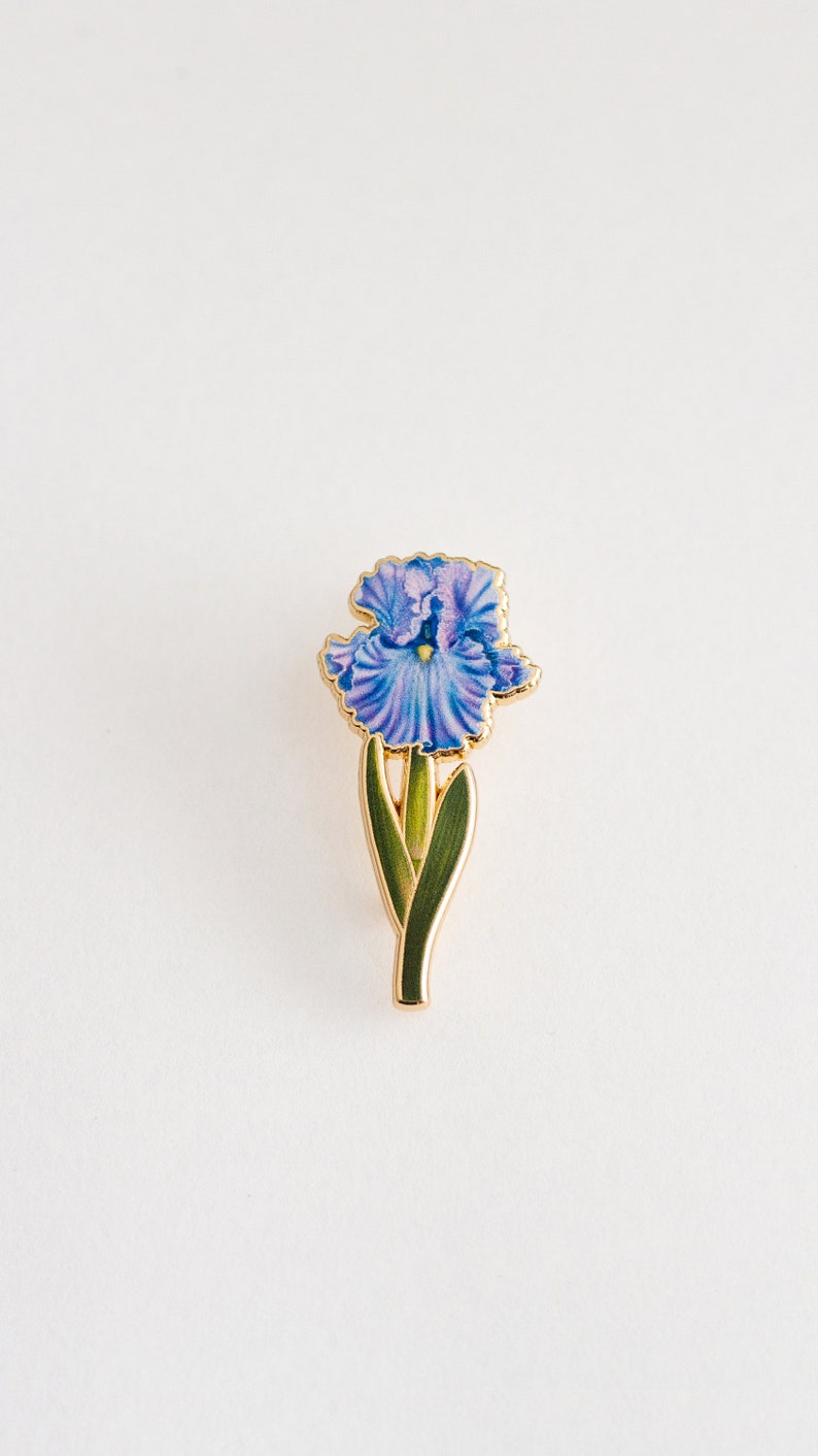 May include: A gold-toned enamel pin featuring a purple iris flower with green leaves.