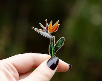 May include: A gold-tone enamel pin featuring a bird of paradise flower with orange, yellow, red, blue, and green accents.