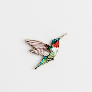 Op de afbeelding: Een kleurrijke kolibrie-speld met uitgespreide vleugels. De vogel heeft een rode keel, een groene rug en blauwe en groene staartveren. De vleugels zijn een mix van roze en wit. De speld staat tegen een witte achtergrond.