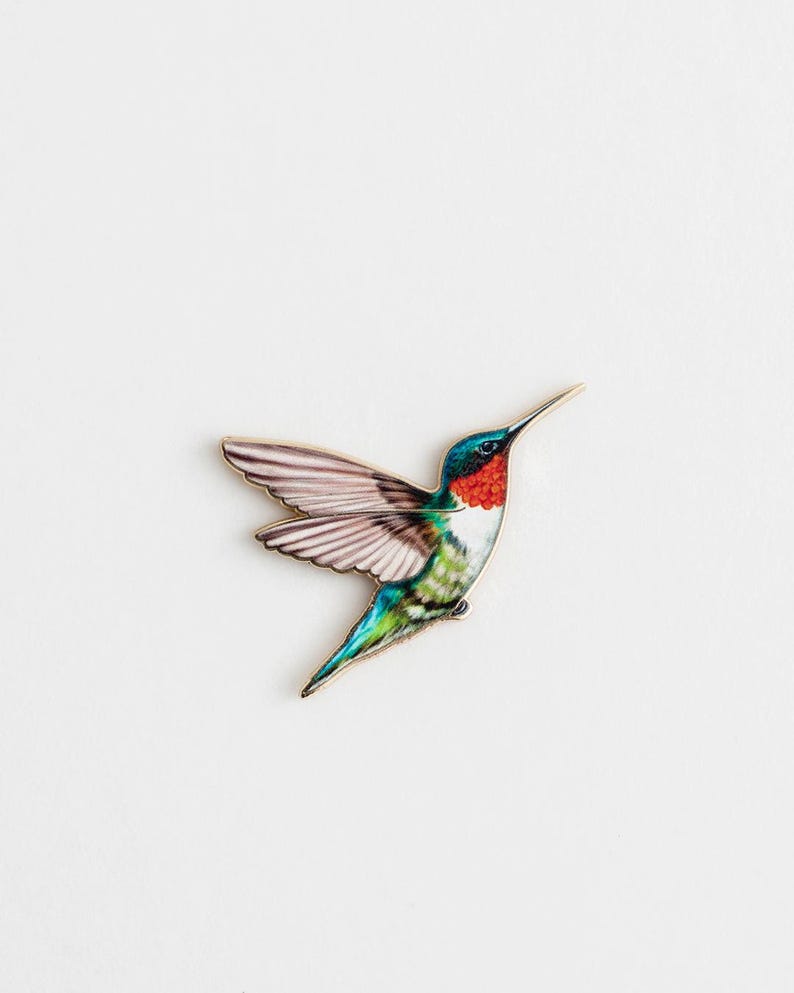 Op de afbeelding: Een kleurrijke kolibrie-speld met uitgespreide vleugels. De vogel heeft een rode keel, een groene rug en blauwe en groene staartveren. De vleugels zijn een mix van roze en wit. De speld staat tegen een witte achtergrond.