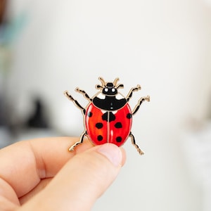 May include: A ladybug enamel pin with a red body, black spots, and a black and white head. The pin has gold-colored metal detailing for the legs and antennae. The ladybug is a popular insect motif.